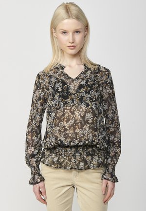 Blouse noire à motifs floraux avec manches longues, décolleté en V, ourlet et poignets volants, fabriquée dans un matériau transparent avec des motifs botaniques en or et blanc détaillés.