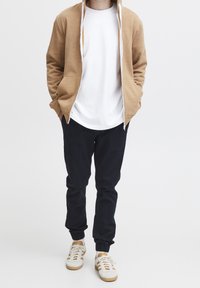 Felpa con zip marrone chiaro, t-shirt bianca, pantaloni jogger neri e sneakers crema con dettagli gialli. Design semplice, vestibilità casual, tessuto testurizzato.