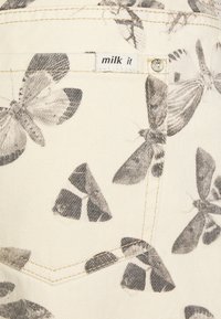 Tecido de denim bege com impressão de borboletas em monocromático, apresentando um bolso traseiro com uma pequena etiqueta "milk it" e um detalhe metálico próximo ao bolso.