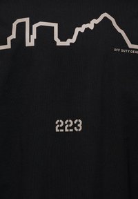 Tissu noir avec une silhouette minimaliste de ville blanche et un contour de montagne, texte « OFF DUTY GEAR » près du sommet et « 223 » en gras centré en dessous.