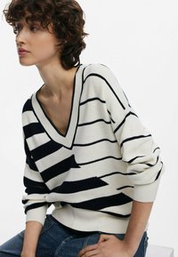 Maglione a righe con scollo a V in crema e blu navy, realizzato in tessuto lavorato a maglia. Caratterizzato da maniche ampie, polsini a coste e una vestibilità rilassata.
