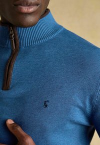 Zils adījuma džemperis ar rāvējslēdzēju kakla daļā, piegriezts ar ribu apkakli un brūniem akcentiem. Neliels izšūts logo krūšu daļā. Gluda tekstūra.