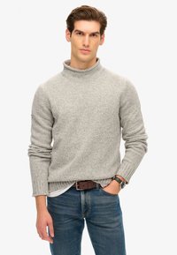 Helles graues Sweatshirt mit hohem Kragen, gestrickter Textur und Bündchen mit Rippenmuster, kombiniert mit blauen Jeans und einem braunen Gürtel.