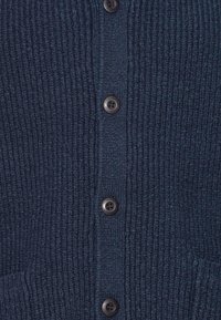 Close-up van een donkerblauwe gestructureerde gebreide cardigan met vier zwarte knopen die verticaal zijn uitgelijnd.