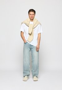 Weißes T-Shirt, hellblaue, verwaschene Jeans, beiger Pullover, um den Hals gebunden, und beiger Slipper, vor einem schlichten Hintergrund präsentiert.