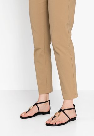 Zwarte platte sandalen met een T-band ontwerp, gouden hardware accenten en een gladde textuur. Gecombineerd met strak aansluitende beige broeken.