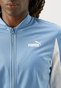 Modrá športová bunda vyrobená z hladkej tkaniny, so stredným zipsom a bielym logom Puma na ľavej strane, s kontrastnými šedými akcentmi.