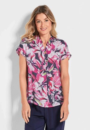 Vrouw die glimlacht, in een kortleurige blouse met grote roze en witte bloemenprint, staande tegen een effen lichte achtergrond.