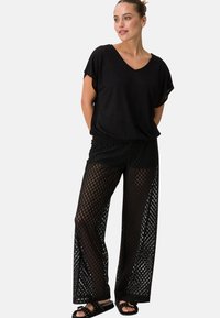 Top nero in maglia con scollo a V, abbinato a pantaloni neri a gamba larga con motivo a griglia e ritagli laterali. Sandali con borchie.