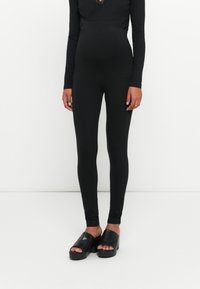 Haut noir à manches longues avec garniture en dentelle associé à des leggings noirs. Le mannequin porte des chaussures à plateforme ouvertes noires avec une finition lisse.