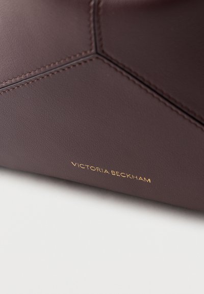 Primer plano de cuero marrón oscuro con costuras hexagonales y el logo dorado de Victoria Beckham grabado cerca del borde.