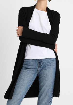 Frau trägt einen langen schwarzen Cardigan, ein weißes T-Shirt und hellblaue Jeans, steht mit verschränkten Armen vor einem einfarbigen Hintergrund.