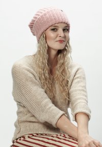 Różowa dzianinowa czapka, beżowy sweter o teksturze oraz spódnica w paski. Miękkie materiały o luźnym kroju i widocznych szwach w neutralnych kolorach.
