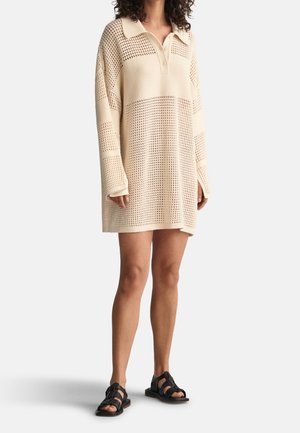 Mujer con un vestido jersey color crema, de punto abierto y oversize, con cuello y botones, combinado con sandalias negras de punta abierta.