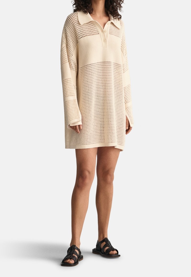Femme portant une robe-pull crème, oversize, en maille ajourée avec un col et des boutons, associée à des sandales noires à bout ouvert.