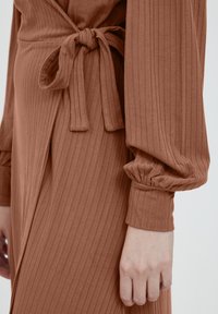 Rostfärgad klänning med ribbad textur, belagd midja och puffärmar. Tyget verkar mjukt och flödande, vilket framhäver dess form.