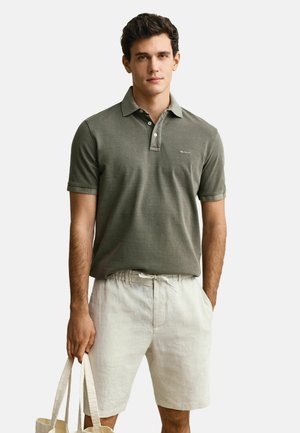 Homme portant un polo vert olive et un short beige clair tenant des sacs fourre-tout beiges sur un fond uni.