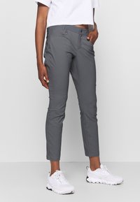 Kobieta w szarych slim-fit spodniach i białych sneakersach, z ręką w kieszeni, na tle jasnym.