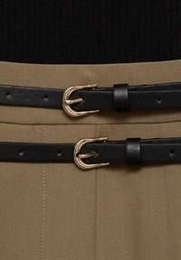 Ceintures en cuir noir avec boucles dorées, superposées sur un tissu beige. La ceinture du bas est légèrement plus large, les deux présentant une texture lisse et des encoches ajustables.