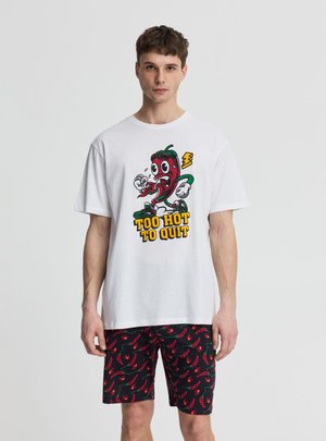 Młody mężczyzna w białej koszuli z nadrukiem kreskówkowej papryczki chili i napisem „Too Hot to Quit”, zestawionej z czarnymi szortami z nadrukiem czerwonych papryczek chili.