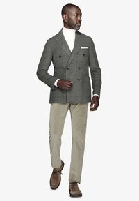 Grå dobbeltbrystet ternet blazer over en hvid turtleneck, parret med beige corduroybukser og brune ruskindssko, stående i profil.