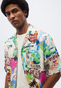 Νεαρός με αφροχτένισμα φοράει πολύχρωμο πουκάμισο με αφηρημένο μοτίβο πάνω από ένα απλό λευκό μπλουζάκι, ποζάρει μπροστά σε απλό φωτεινό φόντο.