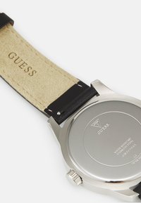 Guess-Uhr mit Edelstahlgehäuse, schwarzem Lederarmband und beige Wildlederfutter. Die Rückseite zeigt eingeprägte Details, einschließlich Markierungen und Logo.