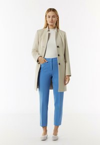 Manteau long beige avec des boutons noirs sur un haut blanc et un pantalon bleu ajusté. Les chaussures sont des ballerines de couleur claire avec un design en maille.