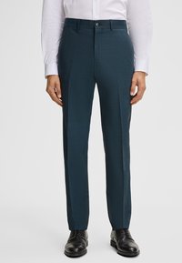 Man in een teal geklede broek, zwarte schoenen en een wit overhemd tegen een effen lichte achtergrond.