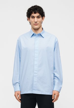 Mężczyzna o kręconych ciemnych włosach, ubrany w jasnoniebieską koszulę z długim rękawem zapinaną na guziki i czarne spodnie, stojący na jednolitym białym tle.