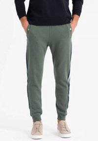 Grüne Jogginghose mit Seitestreifen, aus Baumwollmischmaterial, mit elastischen Bündchen, zwei Vordertaschen und minimalistischem Design. Kombiniert mit beigen Sneakers.