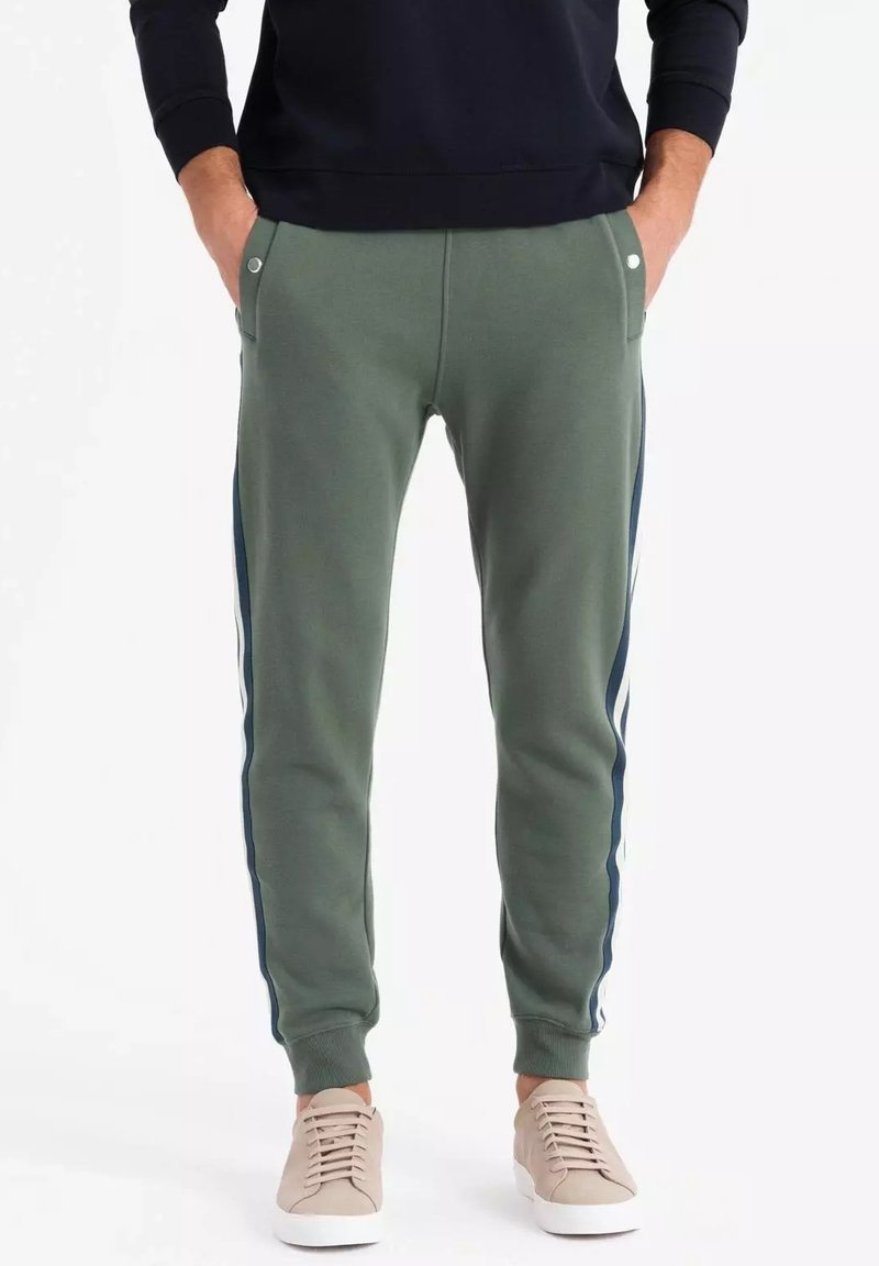 Grüne Jogginghose mit Seitestreifen, aus Baumwollmischmaterial, mit elastischen Bündchen, zwei Vordertaschen und minimalistischem Design. Kombiniert mit beigen Sneakers.