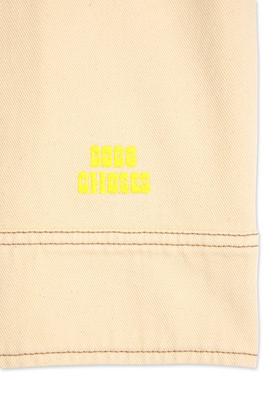 Tessuto di cotone beige con ricamo spesso di colore giallo brillante che recita "BOBO CHOSES" vicino all'orlo inferiore. Texture liscia e design resistente.