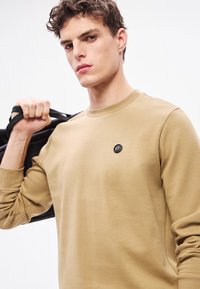 Béžová mikina s okrúhlym výstrihom, na hrudi s čiernym okrúhlym logom. Model drží čiernu tašku cez rameno na jednoduchom pozadí.