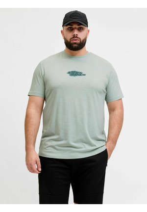 Homme barbu portant un t-shirt vert clair Jack & Jones, un short noir et une casquette noire sur un fond clair uni.