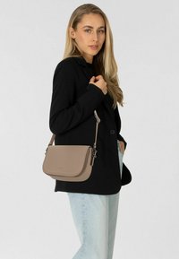 Beige Leder-Crossbody-Tasche mit Stichdetails, einem Klappenverschluss und einem abnehmbaren Tragegurt, getragen zu einem schwarzen Blazer und blauen Jeans.