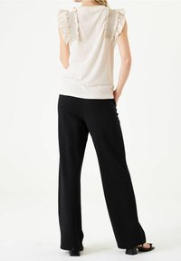 Donna che indossa un top beige senza maniche con spalle arricciate e pantaloni neri a gamba larga, in piedi con una mano in tasca, voltata di spalle.