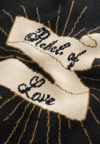 Fekete és bézs szövet egy szalaggall, amelyen a "Szeretetből készült" felirat olvasható díszes írással, arany sugaraival körülvéve egy texturált háttéren.