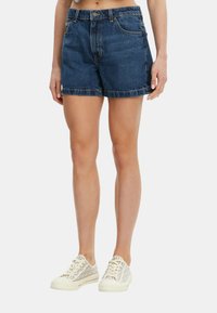 Shorts in denim blu scuro con un design a taglio dritto, caratterizzati da orli sfilacciati e tasche laterali, indossati con sneakers chiare.