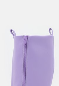 Pochette violette zippée en matériau lisse, dotée de boucles latérales et de coutures contrastantes le long de la fermeture éclair.