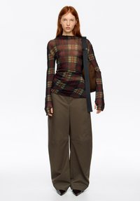 Długi rękaw siateczkowej bluzki w ciemnej kratę z czerwonymi i zielonymi liniami, zestawionej z luźnymi oliwkowymi spodniami i czarnymi szpilkami na spiczastym nosku.