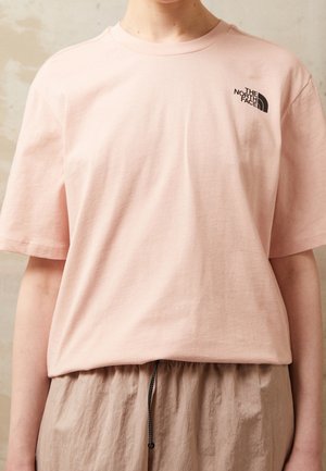 Osoba ubrana w jasnoróżową koszulkę The North Face i beżowe spodnie z czarnym ściągaczem na jednolitym tle.