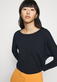 Haut tricot navy à col rond et manches tombantes, associé à un pantalon orange taille haute. Texture lisse, design minimaliste.