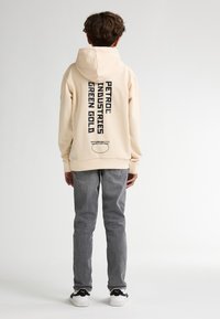 Sudadera crema con un gran logo negro y texto en la parte trasera, combinada con jeans grises y zapatillas en blanco y negro. Textura suave.
