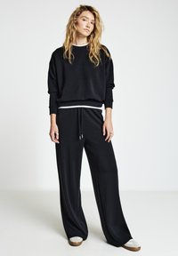 Schwarzer Pullover mit langen Ärmeln und lockerem Sitz, kombiniert mit schwarzen, mit Kordelzug versehenen Hosen. Strukturierter Stoff mit weißem Streifen an der Taille.