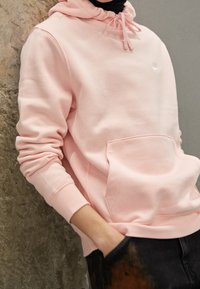 Rosa sweatshirt gjord av mjukt tyg, med en framficka och dragsko-huva. Märkbar logotyp placerad på bröstet. Avslappnad passform.