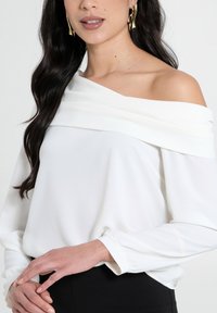 Blusa bianca scollata sulle spalle con maniche lunghe, realizzata in tessuto morbido; presenta un collo drappeggiato e polsini aderenti.
