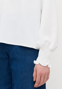 Weißes, strukturiertes Oberteil mit Puffärmeln und Rüschenbündchen. Kombiniert mit blauer Jeans, die eine lockere Passform und einen weichen Stoff zeigt.