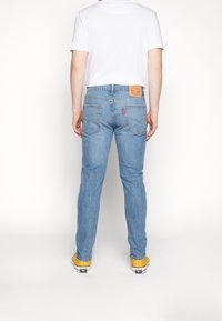 Hombre con jeans de denim azul claro, camiseta blanca y zapatillas amarillas de pie de espaldas contra un fondo blanco liso.