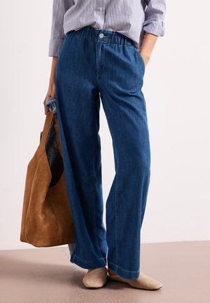 Kvinde iført løstsiddende blå denimbukser og stribet skjorte, der holder en brun ruskindstote, med synlige beige flade sko.